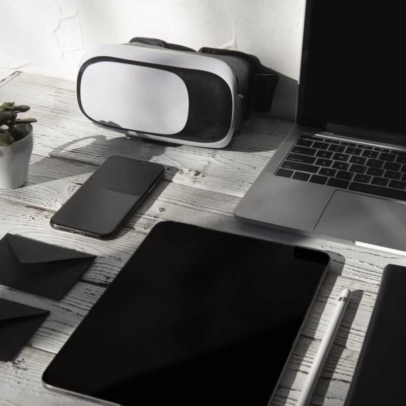 A modern workspace setup featuring a laptop, smartphone, tablet, VR headset, stylus, black envelopes, and a small potted plant on a wooden desk.